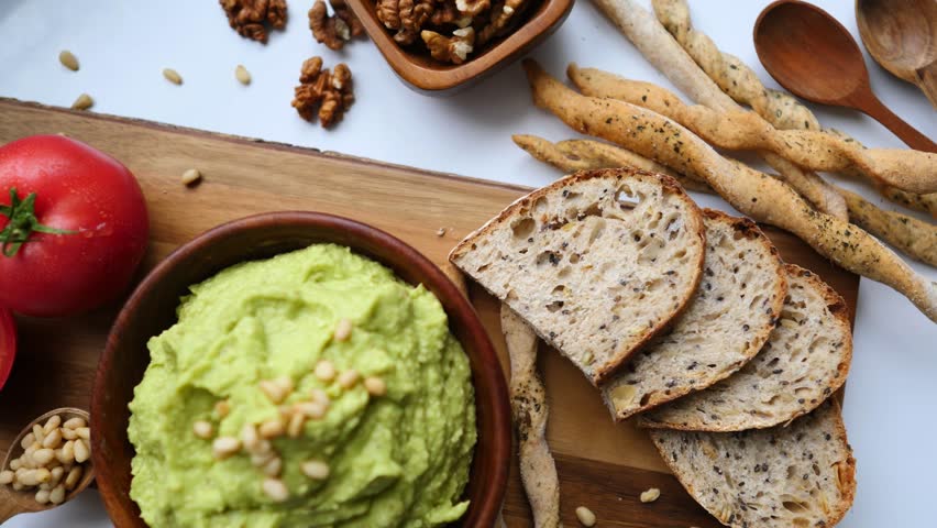 Fresh homemade guacamole in a wooden bowl with pine nuts, served with whole grain bread, breadsticks, and tomatoes. Healthy vegetarian snack or appetizer, perfect for a balanced diet