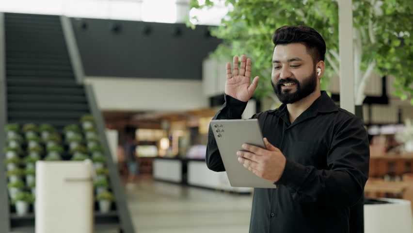 Portrait of friendly Indian salesman with earphones waves hand to interlocutor on tablet in office. South Asian businessman joins video-call via mobile device in shopping mall - Powered by Shutterstock - Get 15% off with code: PIKWIZARD15