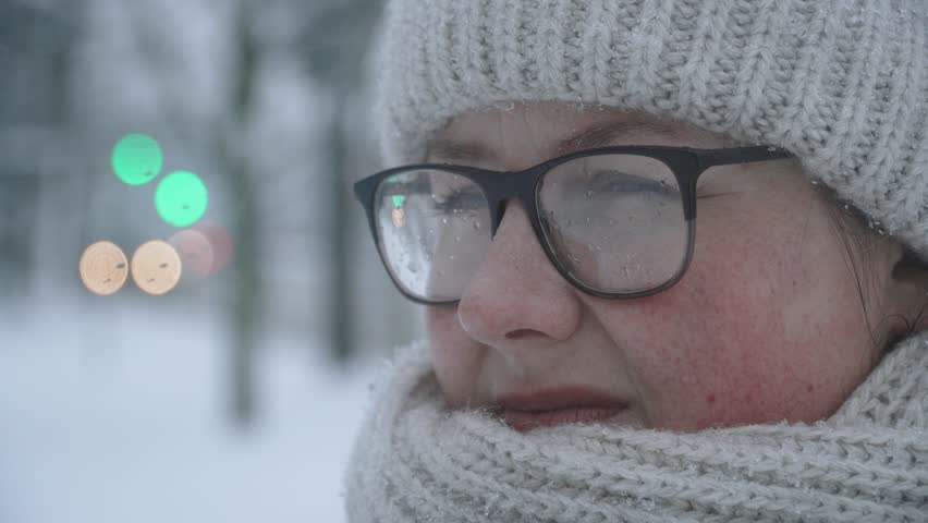 Portrait of distressed young woman trying to keep warm on snowy winter day. Person is waiting outside in snowfall, with melted snowdrops covering her glasses.