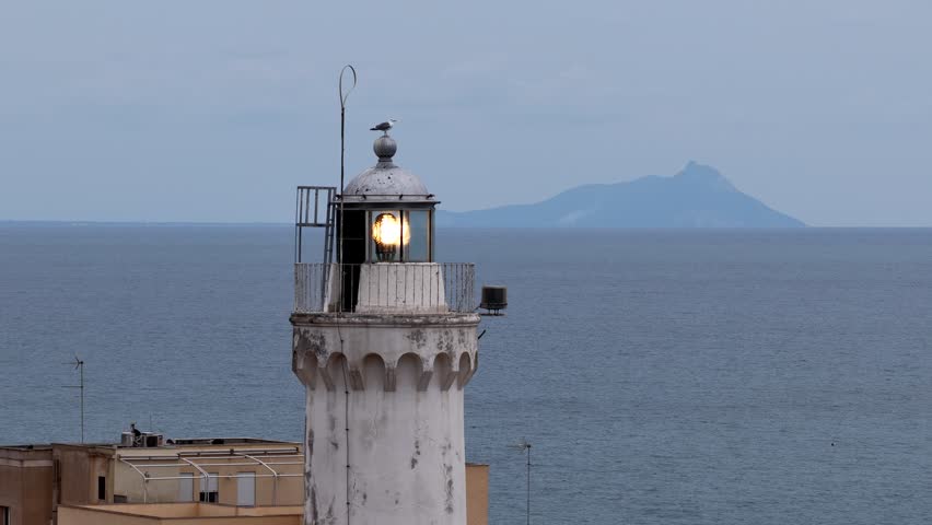 Cinematic aerial close-up of a seagull flying away from the lighthouse on the coast of Anzio, Italy. Peaceful sea view with mountain in the background.
