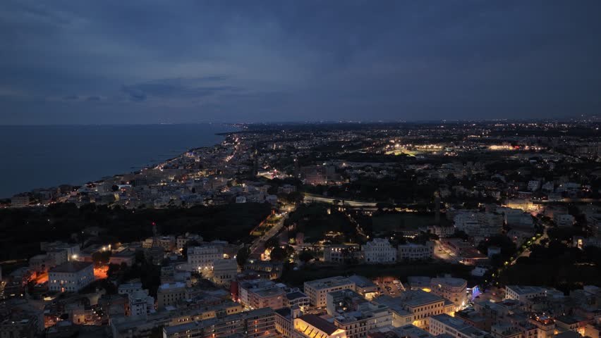 Cinematic drone footage moving backward at dusk over Anzio, Italy. Aerial view of the illuminated harbor and coastline with calm sea and city lights.