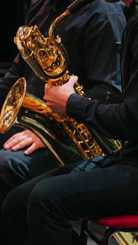 A close-up of two musicians seated on stage. One of them plays a shiny brass baritone saxophone. The orchestra performs