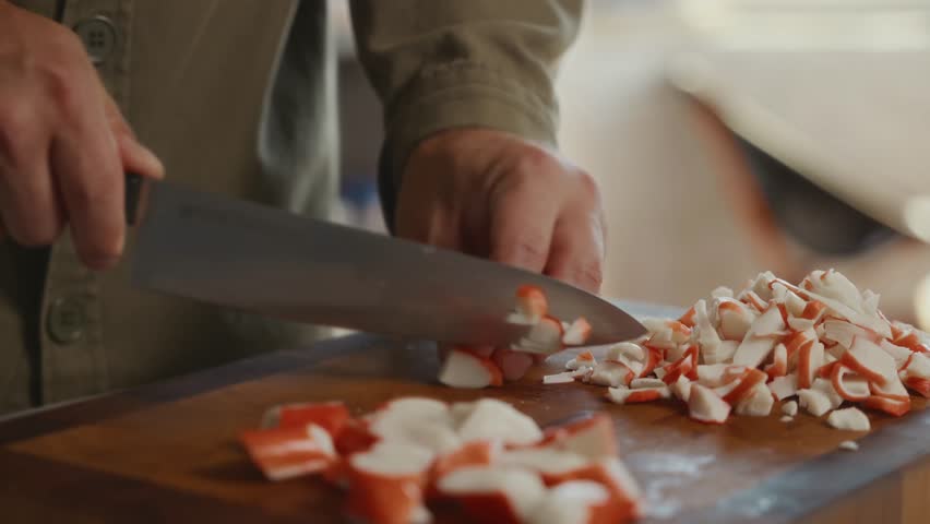 Mans hands chopping crab sticks on wooden board in home kitchen. Cozy minimalist cooking moment reflecting harmony, simplicity, and balance at home. High quality 4k footage