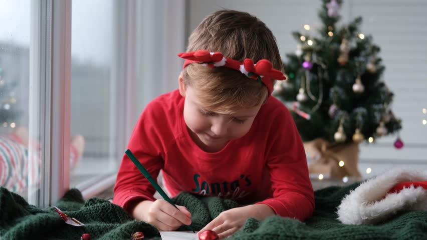 A child in a santa hat draws a picture . Boy on Christmas Eve.