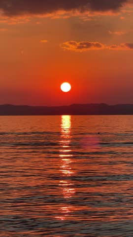 Red-orange sunset over Lake Garda, sun setting behind the horizon, glowing reflection on the water. Dramatic cinematic evening with peaceful lake and warm golden sky in Italy