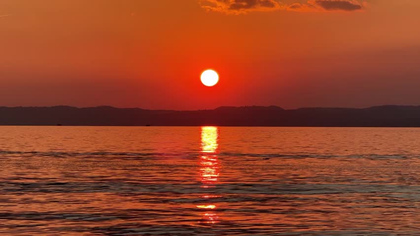 Red-orange sunset over Lake Garda, sun setting behind the horizon, glowing reflection on the water. Dramatic cinematic evening with peaceful lake and warm golden sky in Italy
