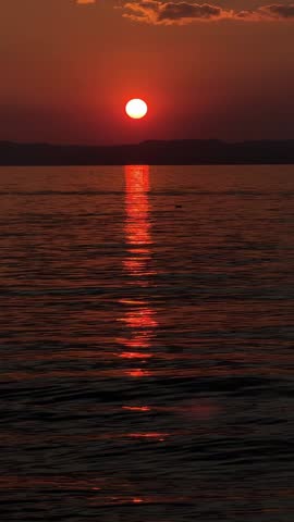 Vivid red-orange sunset over Lake Garda, sun disk dipping below the horizon, reflected on calm water. Cinematic evening scene with dramatic colors over the Italian lake