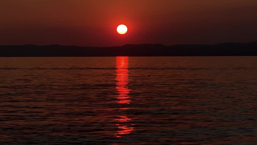 Vivid red-orange sunset over Lake Garda, sun disk dipping below the horizon, reflected on calm water. Cinematic evening scene with dramatic colors over the Italian lake