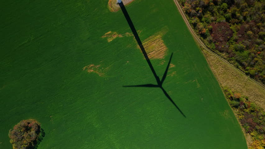Aerial top view of a green field with the shadow of a large wind turbine stretching across the landscape. High quality 4k footage