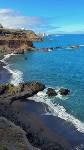 Aerial view of Bollullo black sand beach in Tenerife, surrounded by volcanic cliffs and turquoise ocean waves. People sunbathing and swimming on the wild Atlantic coast