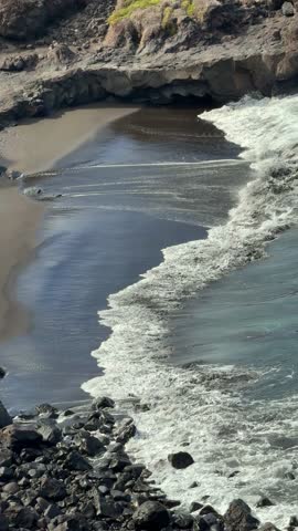 view of Bollullo black sand beach in Tenerife, Spain. Powerful Atlantic waves crash onto volcanic sand surrounded by dramatic lava cliffs on the wild Canary coast