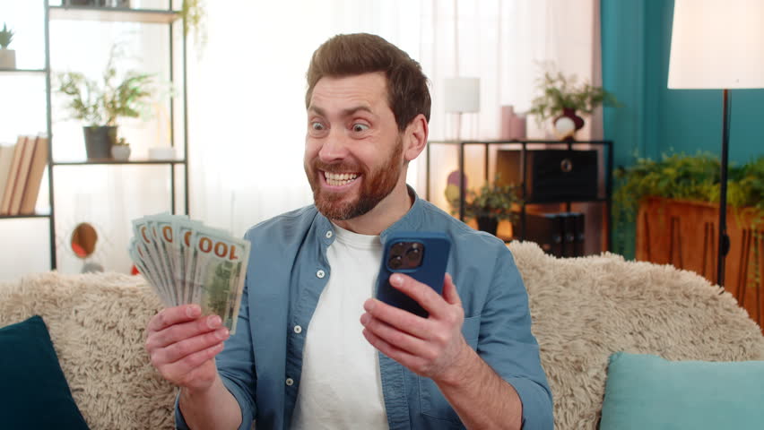 Man at home checks his balance on a smartphone with an excited expression, thrilled about received money. Caucasian guy smiles, celebrating a lottery or online game win, feeling lucky and successful.