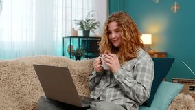 Young woman at home smiling while watching online movie on laptop and drinking hot tea with pleasure. Caucasian redhead girl on sofa enjoying peaceful time, cozy evening, comfort and relaxation - Powered by Shutterstock - Get 15% off with code: PIKWIZARD15