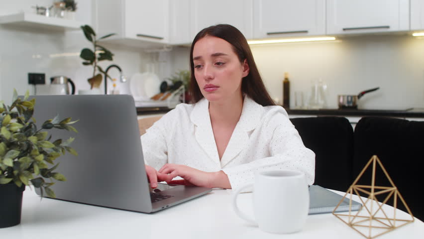 Young woman at home works on laptop leans on hand bored tired low drive feels burnout needs rest. Caucasian girl on sofa sits at table looks at screen slow typing sighs low mood demotivated and dull.
