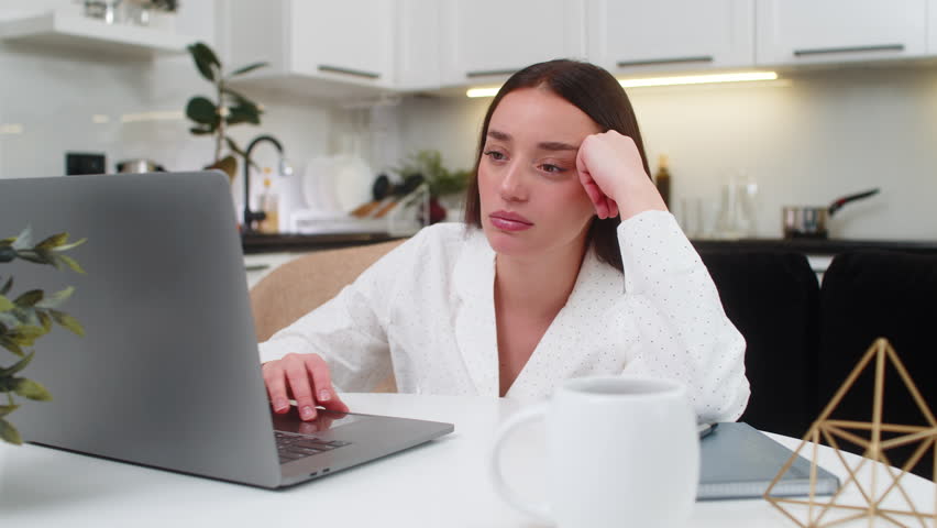 Young woman at home works on laptop leans on hand bored tired low drive feels burnout needs rest. Caucasian girl on sofa sits at table looks at screen slow typing sighs low mood demotivated and dull.