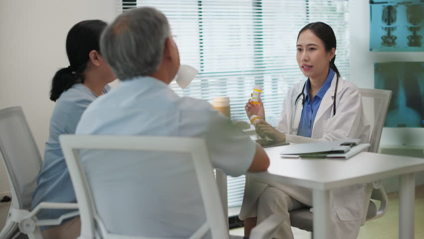 Asian female doctor explains how to take prescription medicine to elderly couple during a medical consultation in a modern clinic. Senior healthcare and patient care at hospital concept.