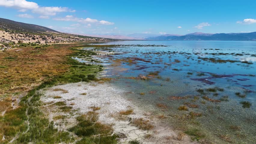 Sweeping aerial drone flight over a scenic lake, revealing a vast shoreline with wetlands, a distant village, and mountains under a blue, cloudy sky