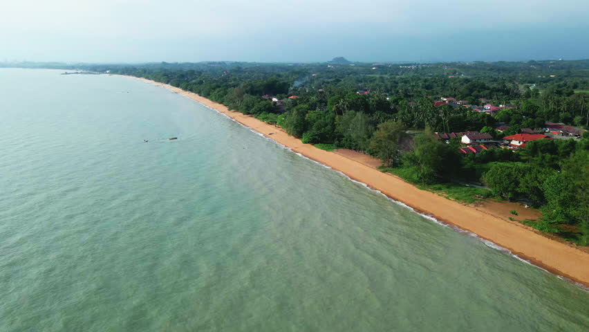 Aerial view of a tropical beach coastline in Malaysia with golden sand, calm sea waves, and nearby houses surrounded by lush greenery.