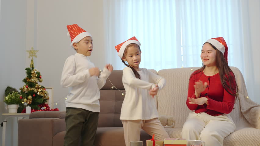 Happy christmas family celebrating holiday at home, parent and two kids enjoy cheerful moment with christmas tree, children dancing enthusiastically, adult parent clapping with presents nearby.