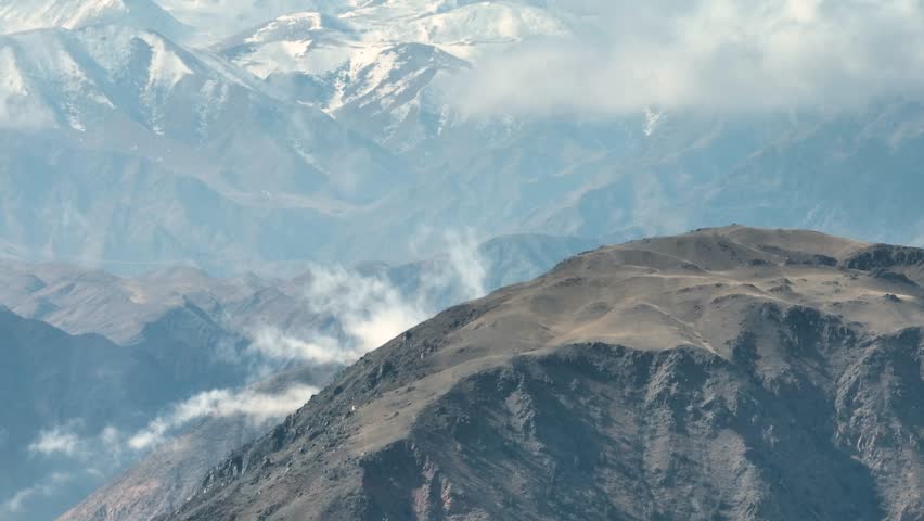 Multiple small plumes of smoke, steam, or dust rise from the slopes of a barren, dark hill, layered against hazy, blue distant mountains and snow capped peaks