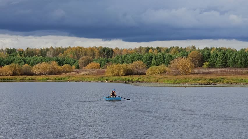 Man is rowing a boat on a lake. The sky is cloudy and the water is calm