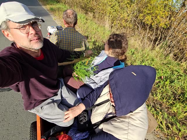 Man is sitting on a cart gasoline bike plow with trailer with a two woman and driver. The man is taking a selfie picture of the scene