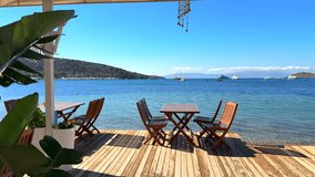 Seaside dining deck with wooden tables and chairs overlooking a beautiful turquoise bay, sailboats, and distant hills under a clear blue sky. - Powered by Shutterstock - Get 15% off with code: PIKWIZARD15