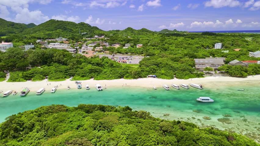 A stunning aerial pan over the crystal-clear turquoise waters of Kabira Bay on Ishigaki Island, Japan. The shot reveals tour boats on a white sand beach surrounded by lush tropical greenery.