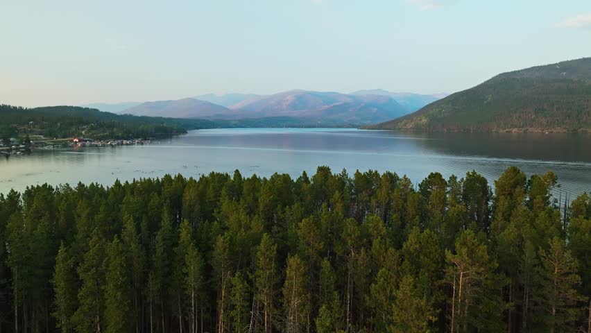 Drone ascend reveal across pine forest and Grand Lake Colorado with calm water and surrounding hills in clear daylight, backdrop