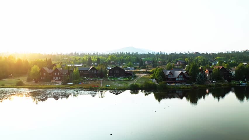 Grand Lake Colorado during sunrise with golden hour reflections across water and distant forest backdrop, aerial pullback from lakeshore lakefront homes