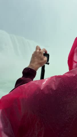 SLOW MOTION SHOT - A tourist in a red poncho onboard a boat filming the approach to Niagara Falls, with mist rising and lush cliffs under a sunny sky, Ontario, Canada.