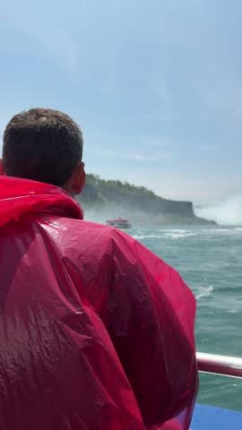 A tourist in a red poncho onboard a boat approaching Niagara Falls, with mist rising and lush cliffs under a sunny sky, Ontario, Canada.