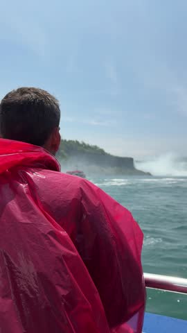 SLOW MOTION SHOT - A tourist in a red poncho onboard a boat approaching Niagara Falls, with mist rising and lush cliffs under a sunny sky, Ontario, Canada.