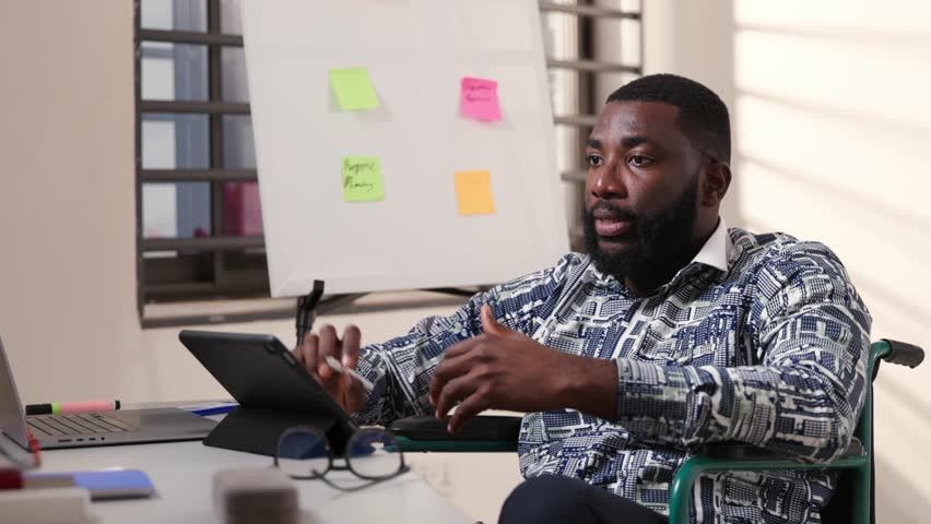 African male corporate professional in a wheelchair uses tablet for remote work, video calls, online shopping, digital banking, social media internet browsing in the office - Powered by Shutterstock - Get 15% off with code: PIKWIZARD15