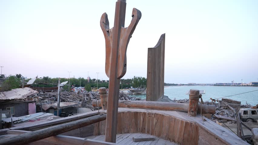 A view from the deck of a traditional wooden boat anchored near a shore, surrounded by remnants of previous vessels. The water reflects the sky, adding to the tranquil atmosphere.