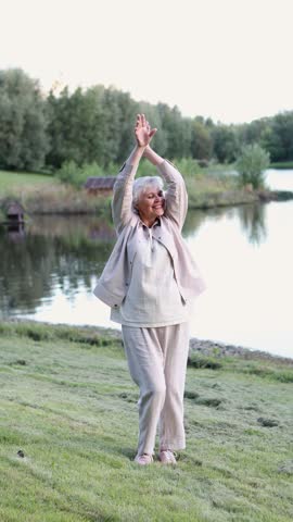 Happy senior woman dancing in nature near scenic lake in park. Energetic retired female enjoying health and positive emotions outdoors, celebrating life and beauty of aging in summer landscape