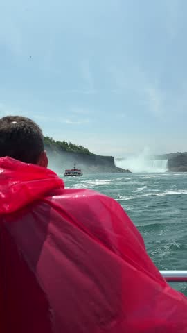 SLOW MOTION SHOT - A tourist in a red poncho onboard a boat approaching Niagara Falls, with mist rising and lush cliffs under a sunny sky, Ontario, Canada.