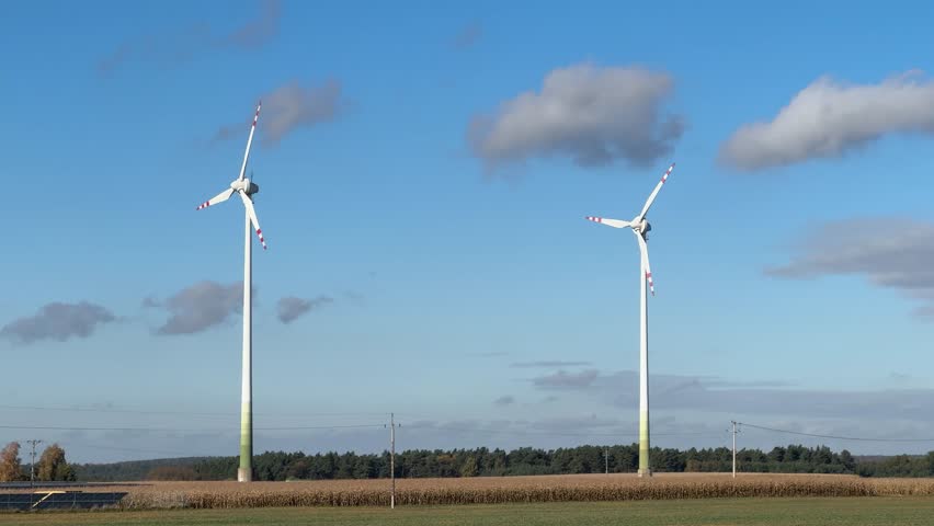 A cinematic hero shot of wind turbines standing tall in a wide green field beneath a clear blue sky with drifting clouds, representing clean renewable energy and environmental technology.