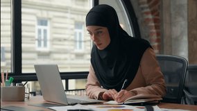 Ethnic concentrated businesswoman Muslim girl business woman Arabian female employer entrepreneur working at table in office with documents papers writing notes reviewing diagram stats using laptop - Powered by Shutterstock - Get 15% off with code: PIKWIZARD15
