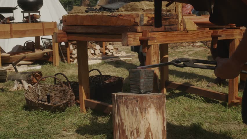 Blacksmith hammering hot iron on anvil with large hammer, side view on a sunny day