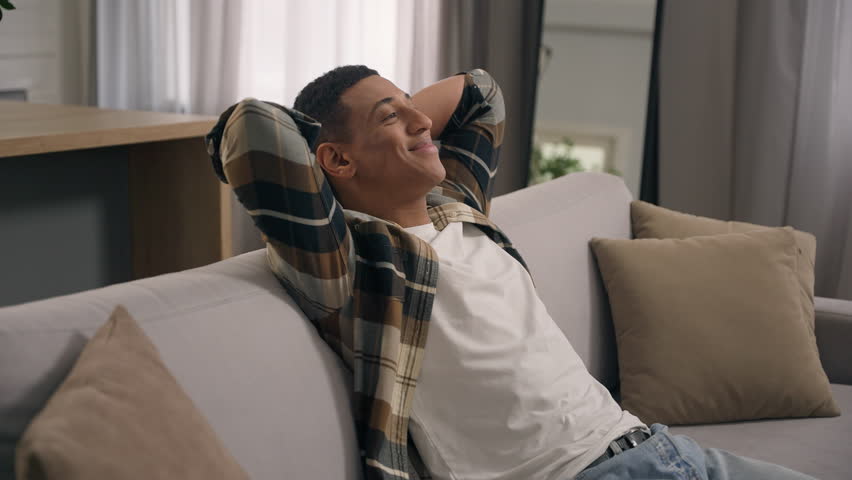 Smiling resting calm African American man guy ethnic male enjoy air conditioner indoors relaxing at home sofa couch leaning back holding hands behind head homeowner napping closed eyes loan mortgage