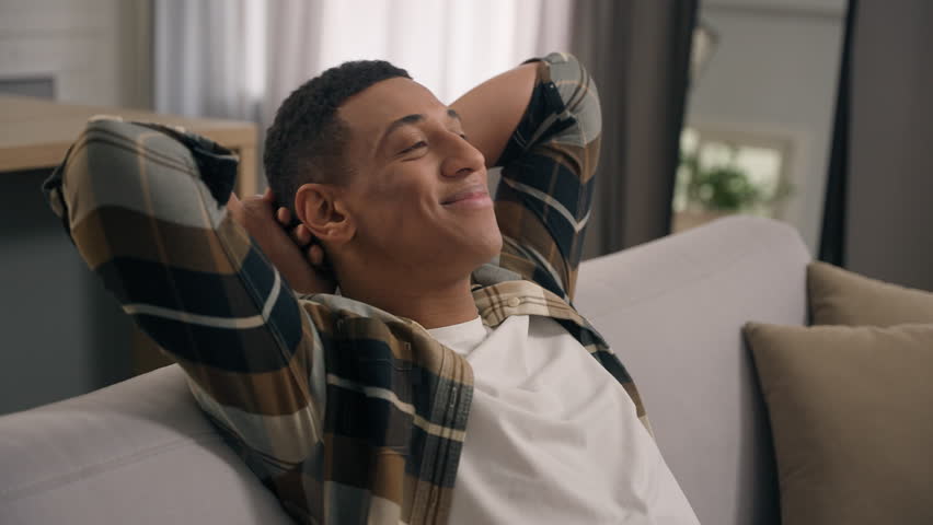 Smiling resting calm African American man guy ethnic male enjoy air conditioner indoors relaxing at home sofa couch leaning back holding hands behind head homeowner napping closed eyes loan mortgage