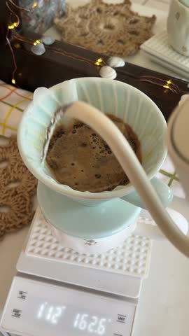 Cozy pour-over coffee brewing setup with warm candlelight and natural decor. Fresh hot water poured into filter over glass server on scale. Relaxing home barista morning scene.
