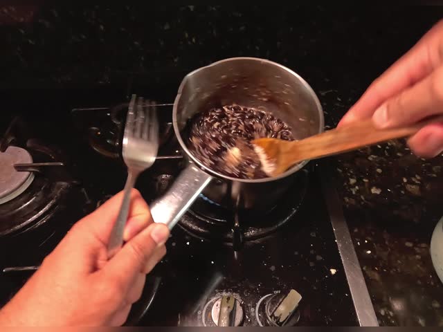 Point of view of male hands stirring beans with cheese and serving them on a plate on a black kitchen counter with black marble and low lighting with yellow lighting.