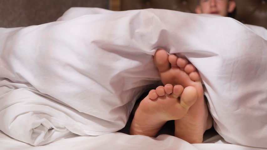 Close-up of bare feet of a boy sticking out from under the blanket and rubbing against each other to scratch. The boy hides his legs back under the blanket. Morning awakening