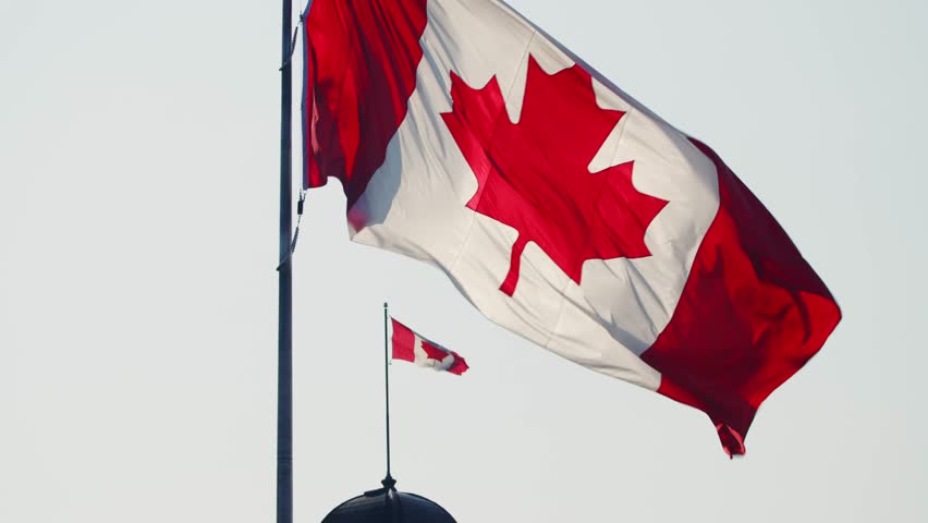 Beautiful View Of The Large Canadian Flag Waving In Warm Sunset Light In Downtown Halifax Nova Scotia Canada, With A Smaller Flag On A Tower In The Background Against The Golden Sky.