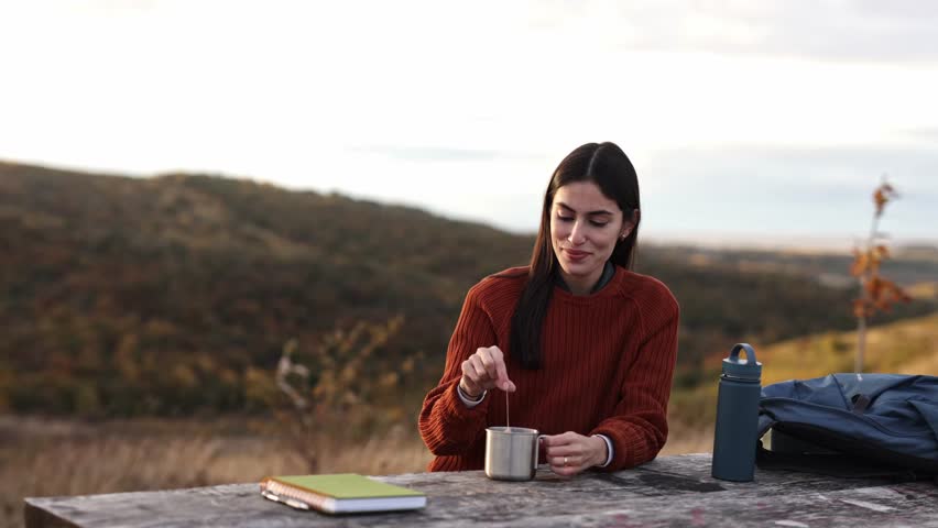 Woman smiling, holding a steaming mug, relaxing outdoors with a backpack during a pleasant autumn hiking trip. 4k video