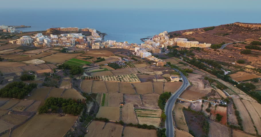 Malta, Comino island: Cultivated fields and a coastal road glowing in sunset light create a tranquil aerial view of Marsalforn, Gozo, Malta. Aerial drone flight footage