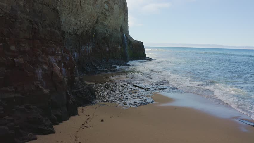 Waterfall cascades from cliff into the Pacific Ocean at Davenport Pier Beach along Highway 1, California, USA. Scenic coastal view with waves, rocky shoreline and blue sky