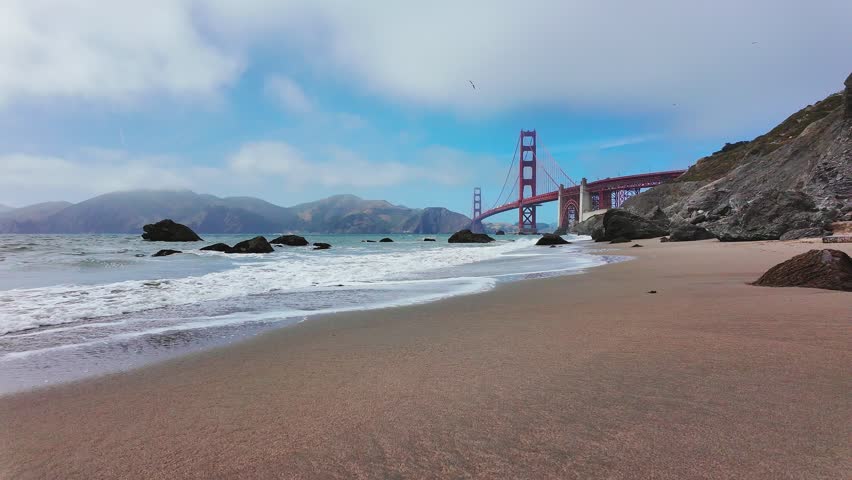 Waves roll onto sandy shore at Baker Beach with Golden Gate Bridge in background, San Francisco, California, USA. Iconic coastal view with ocean, rocks and Marin Headlands